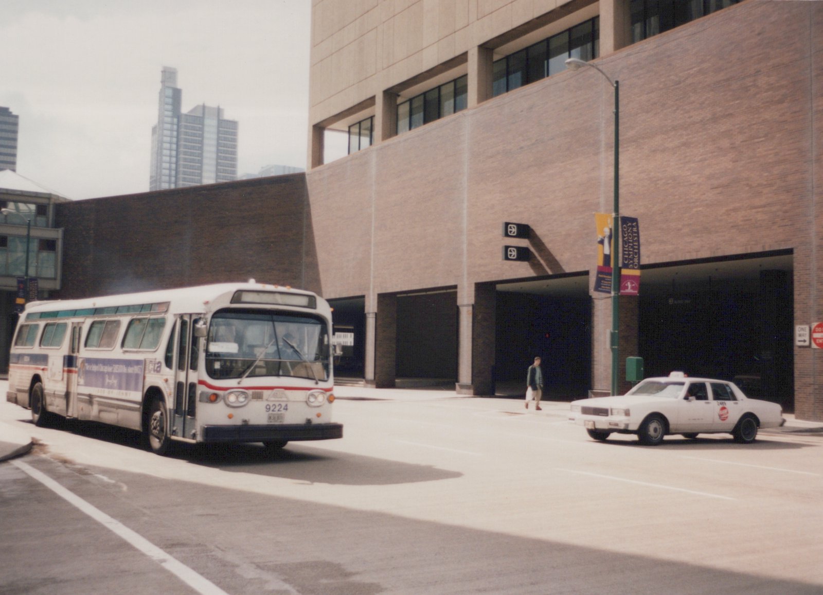 A white and blue CTA bus drives down a city street, with a tan building and a white taxi cab in the background.
