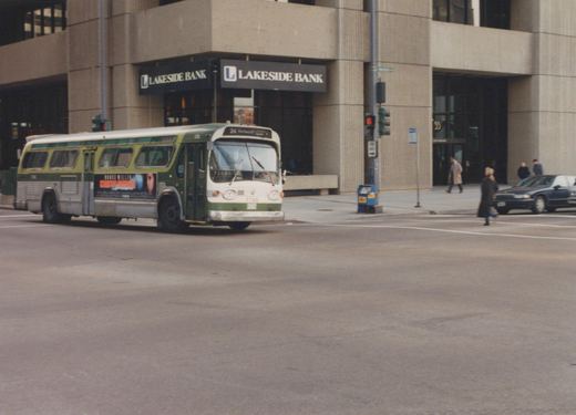 A green and cream CTA bus is stopped at an intersection in front of a building with a Lakeside Bank sign, with pedestrians crossing the street.