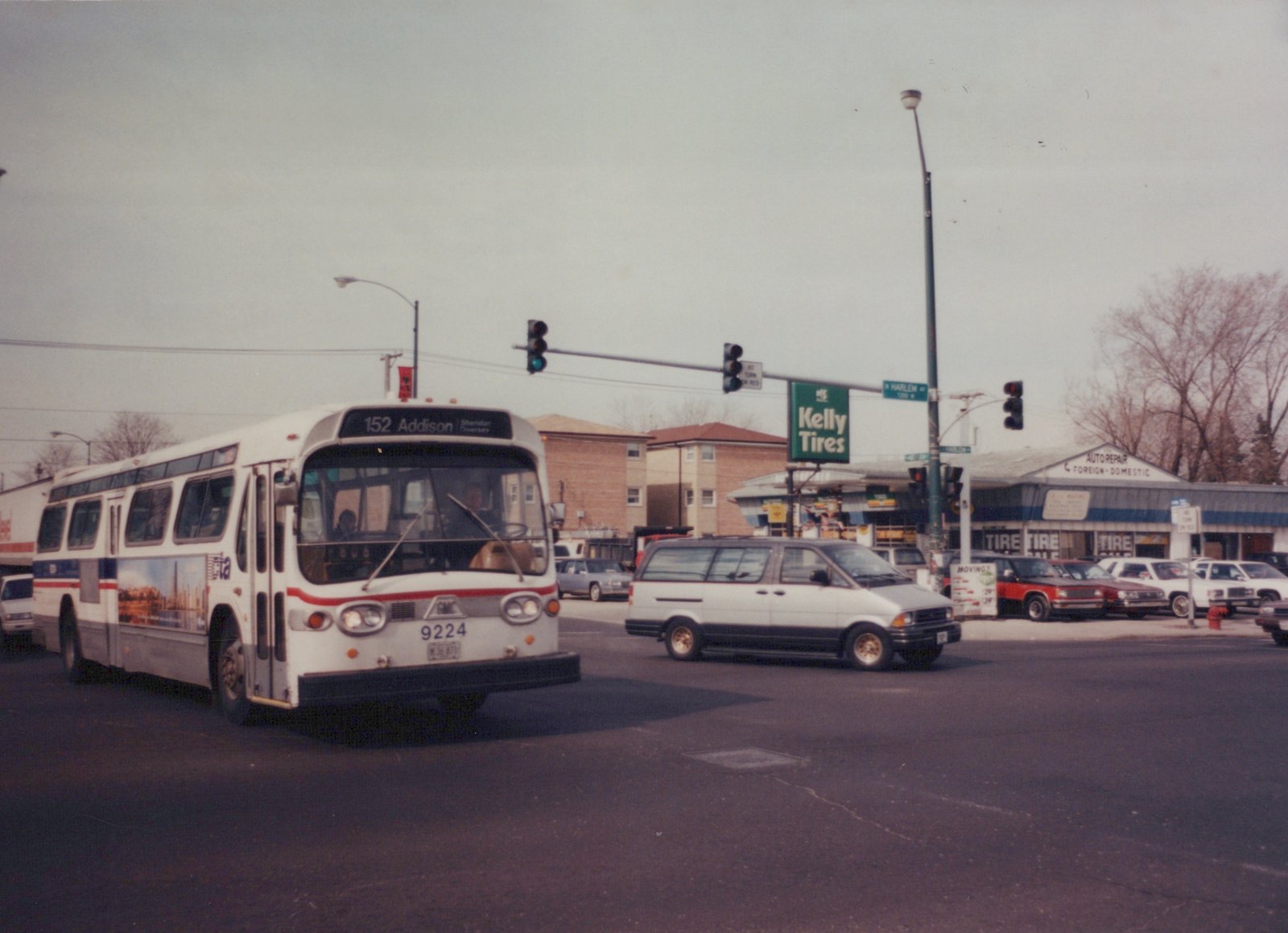 A white Chicago Transit Authority bus with a red and blue stripe, displaying route 152 Addison, is turning at an intersection with a Kelly Tires sign and bare trees in the background.