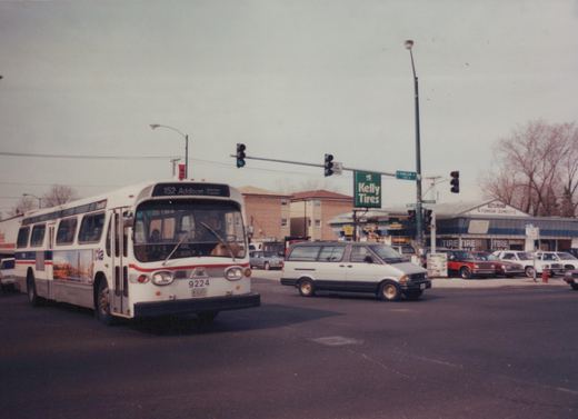 A white Chicago Transit Authority bus with a red and blue stripe, displaying route 152 Addison, is turning at an intersection with a Kelly Tires sign and bare trees in the background.