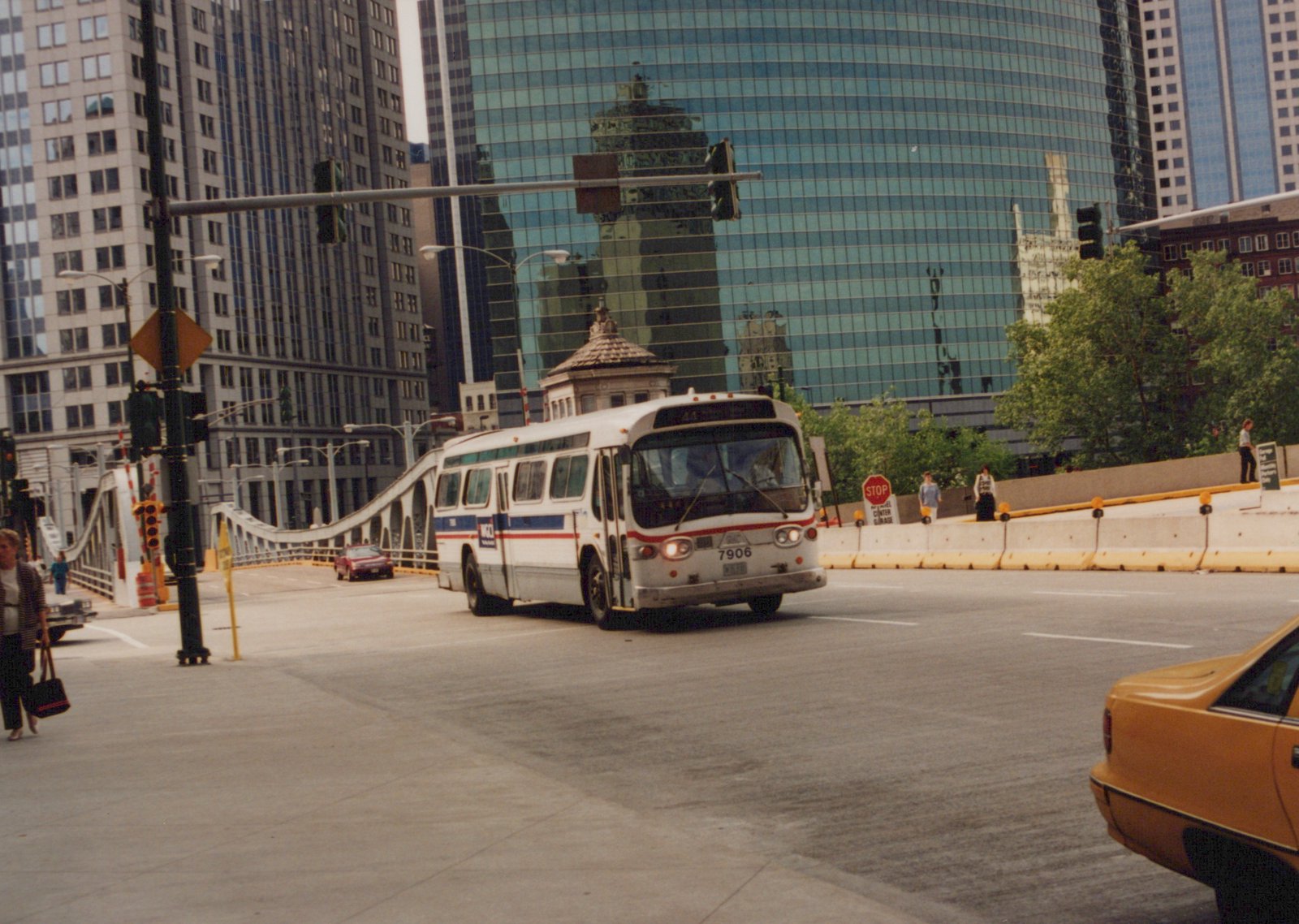 A white CTA bus with the number 7906 drives on a city street with tall buildings in the background and pedestrians on the sidewalk.