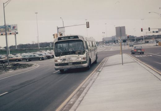 A white and green Chicago Transit Authority bus, number 7934, on route 40 to O'Hare, is driving on a multi-lane road with other cars and a large billboard in the background on a cloudy day.