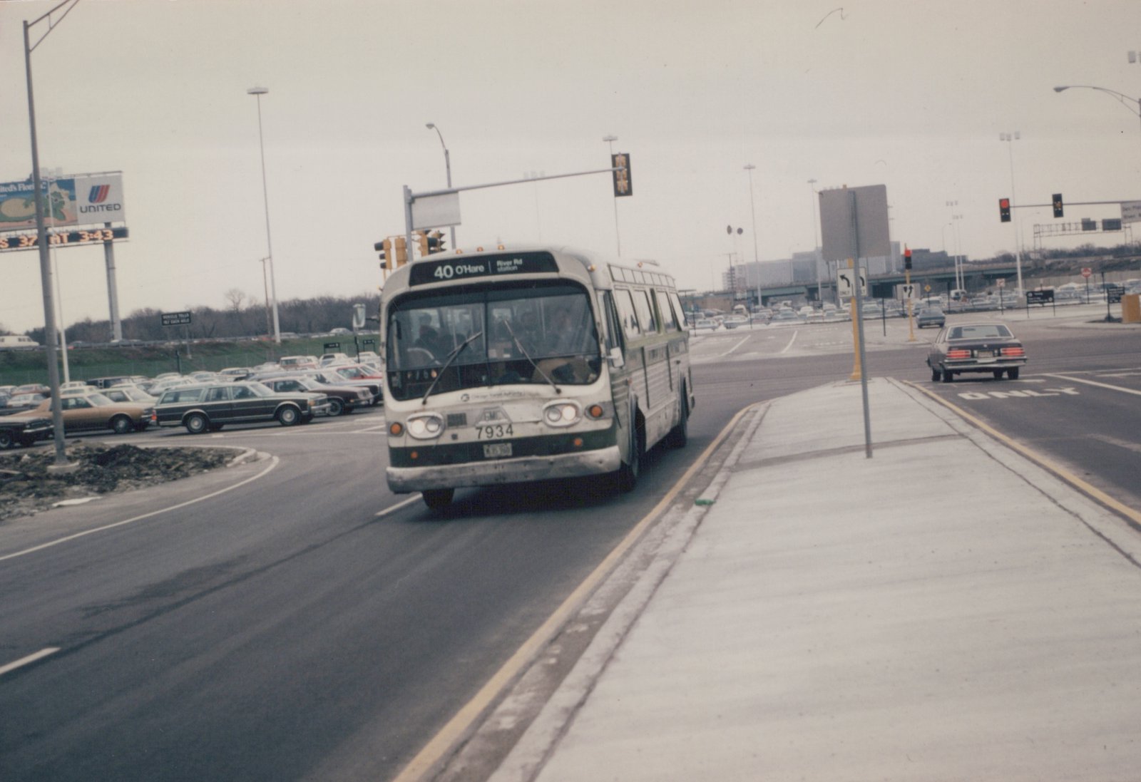 A white and green Chicago Transit Authority bus, number 7934, on route 40 to O'Hare, is driving on a multi-lane road with other cars and a large billboard in the background on a cloudy day.
