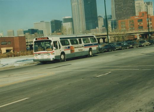 A white and blue Chicago Transit Authority bus drives down a wide city street on a clear winter day, with tall buildings and parked cars behind it.