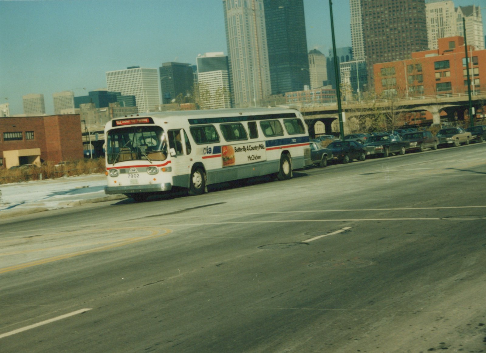 A white and blue Chicago Transit Authority bus drives down a wide city street on a clear winter day, with tall buildings and parked cars behind it.