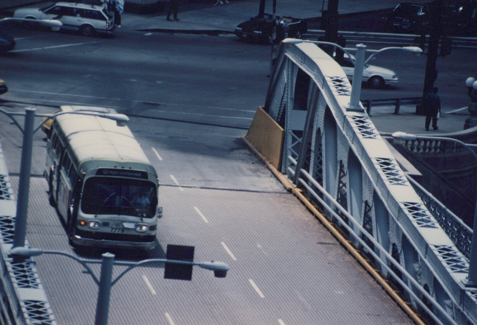 An elevated view of a green and cream Chicago Transit Authority motor bus, number 7779, driving on a bridge over a street with cars and a pedestrian visible below.