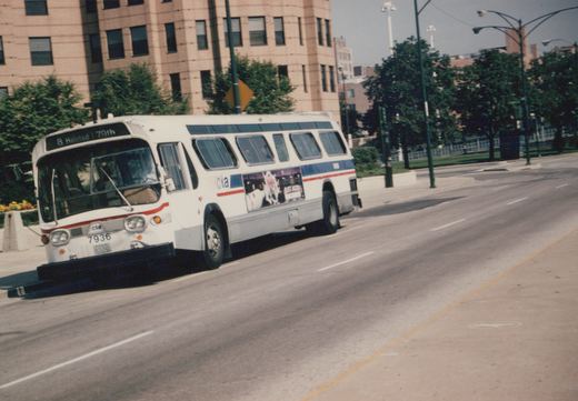 A white Chicago Transit Authority bus with blue and red stripes, numbered 7936, drives down a city street on a clear day.