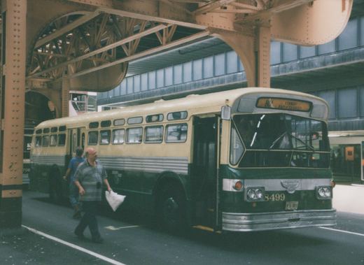 A cream and green CTA bus numbered 8499 is stopped under a steel bridge truss with two men walking nearby.