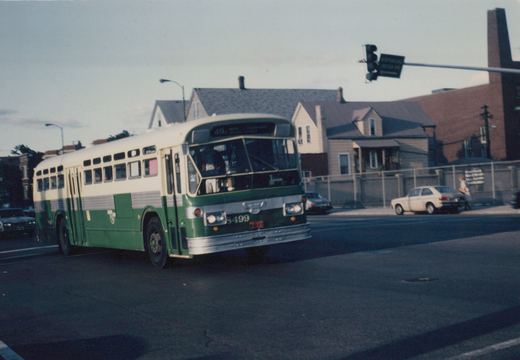 A green and cream CTA bus with the number 8499 on its front is stopped at a street intersection with houses and a traffic light in the background.