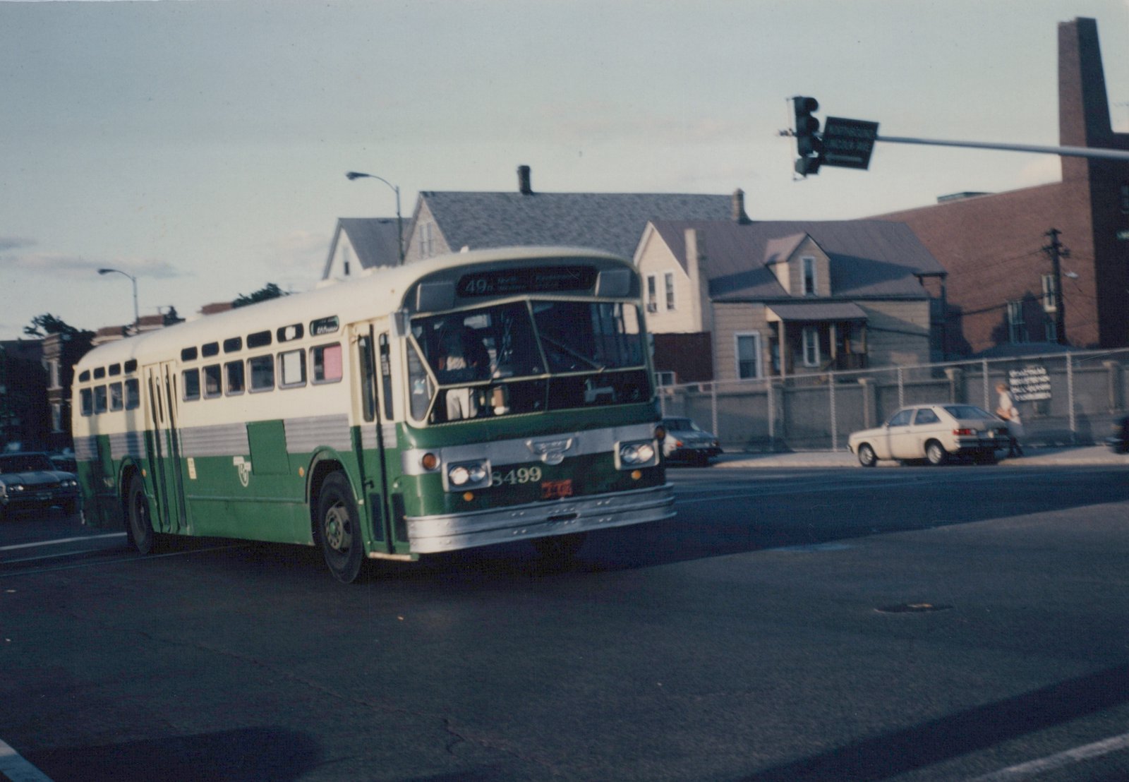 A green and cream CTA bus with the number 8499 on its front is stopped at a street intersection with houses and a traffic light in the background.