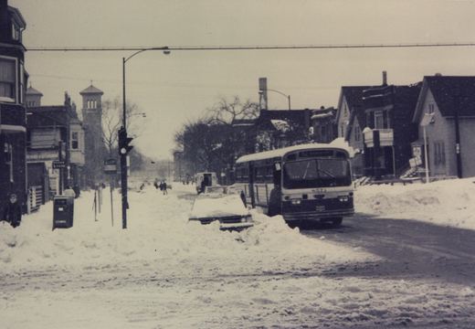 A black and white photo of a CTA bus driving down a snow-covered street with buildings and pedestrians in the background.
