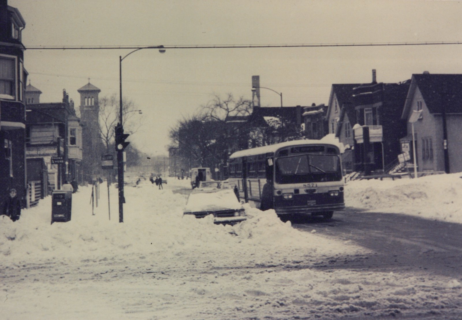 A black and white photo of a CTA bus driving down a snow-covered street with buildings and pedestrians in the background.