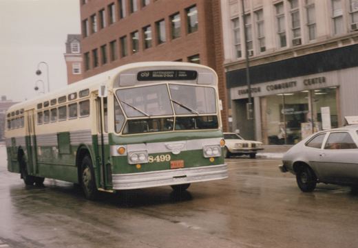 A green and cream Flxible New Look bus numbered 8499, operated by Northeastern Illinois U-bus, is shown on a wet street with a sign for Evanston Community Center in the background.