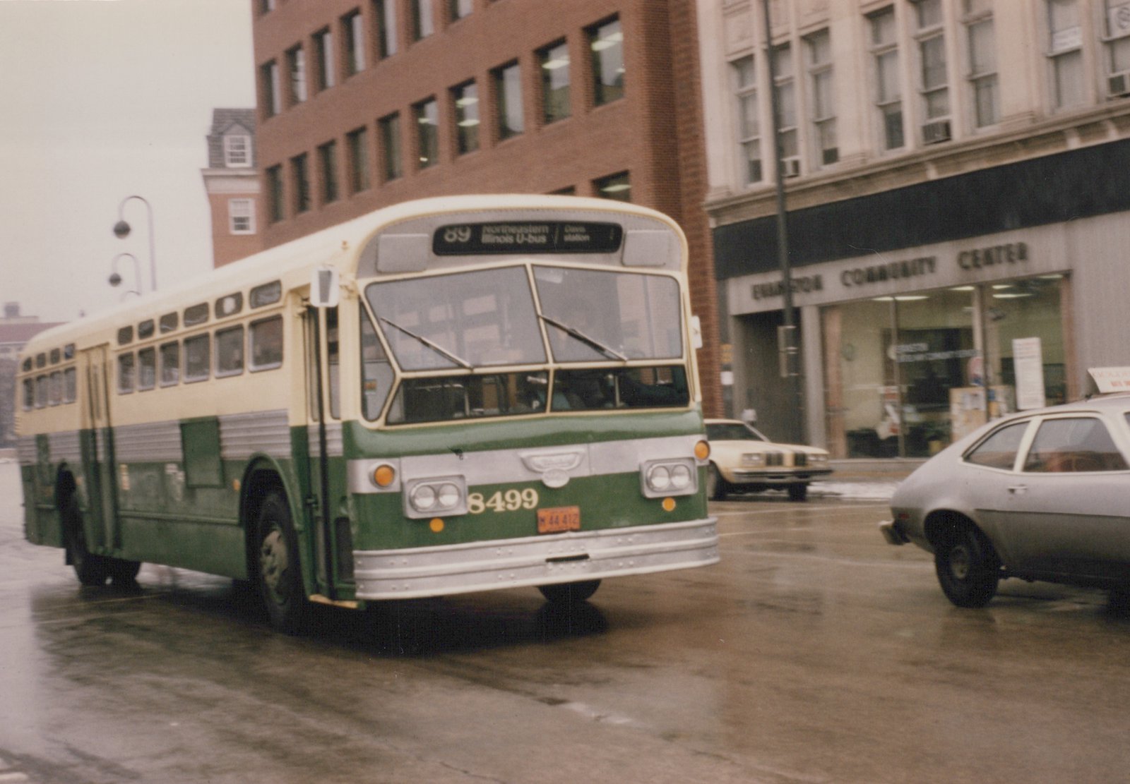 A green and cream Flxible New Look bus numbered 8499, operated by Northeastern Illinois U-bus, is shown on a wet street with a sign for Evanston Community Center in the background.