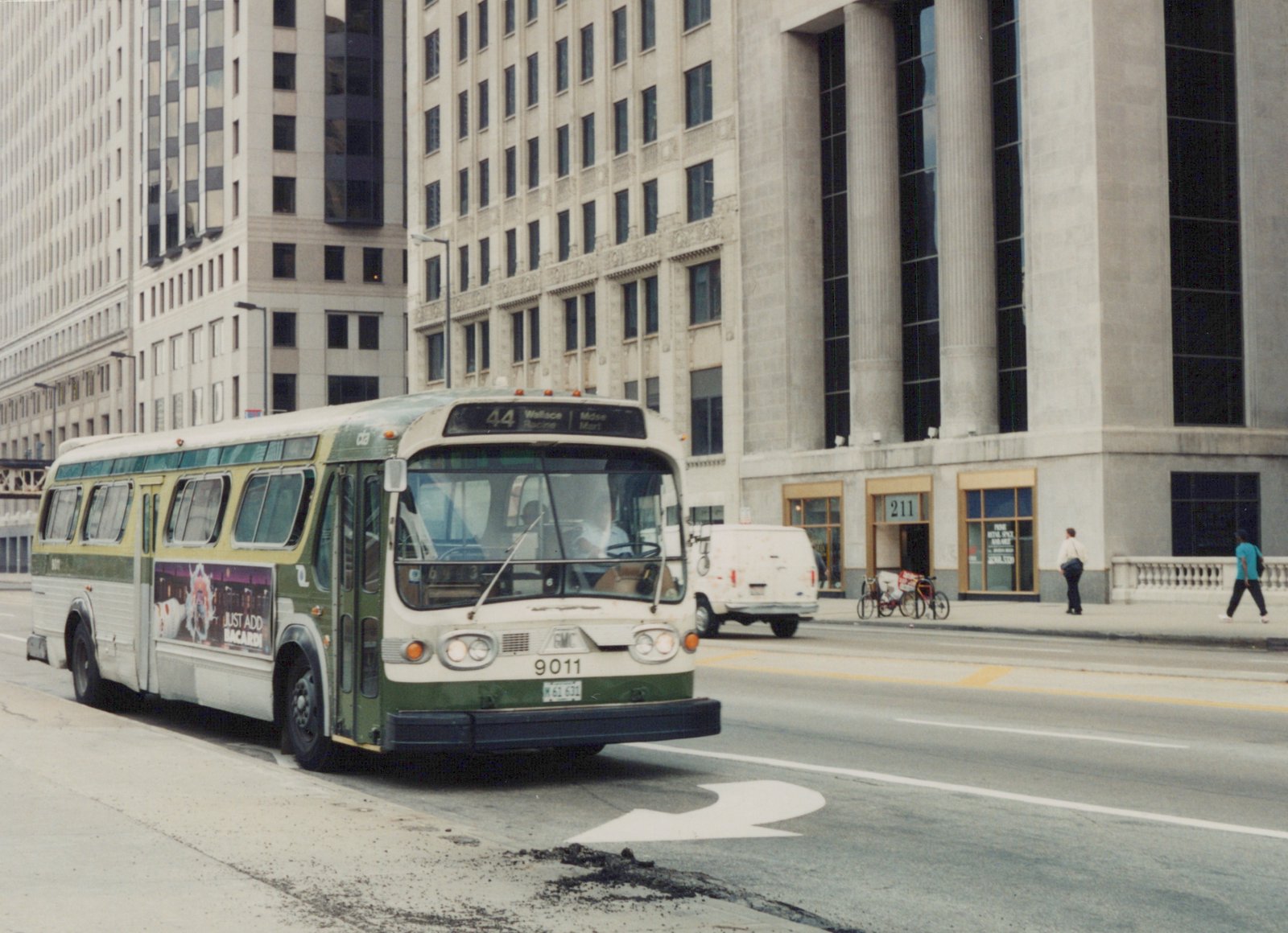 A green and cream CTA bus on route 44 is turning on a city street in front of large stone buildings, with pedestrians on the sidewalk.