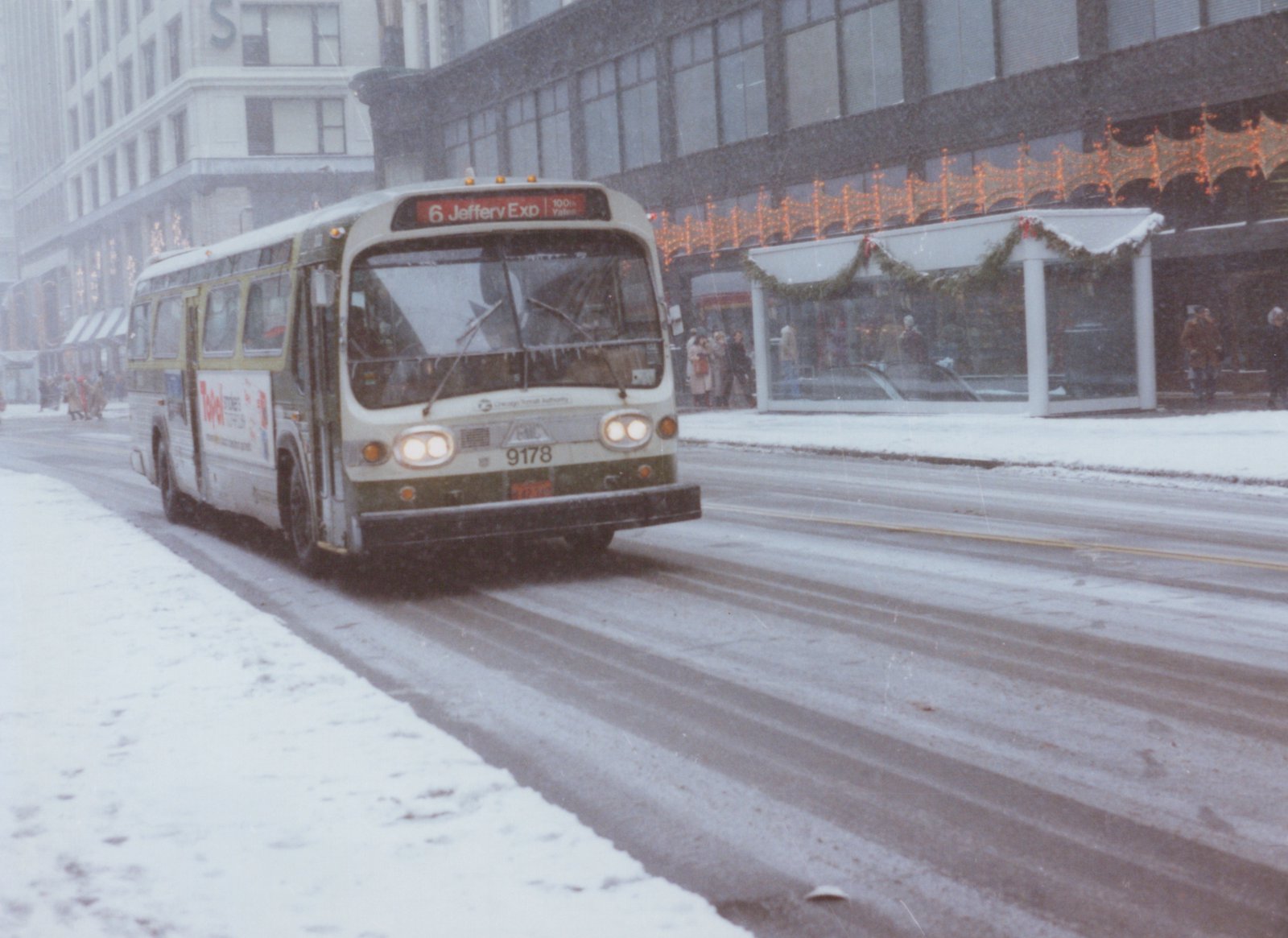 A white and green Chicago Transit Authority bus, number 9178, drives on a snowy street with Christmas lights on the building behind it.