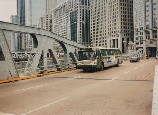 A green and white CTA bus, numbered 9101, drives on a steel bridge in a city with tall buildings in the background.
