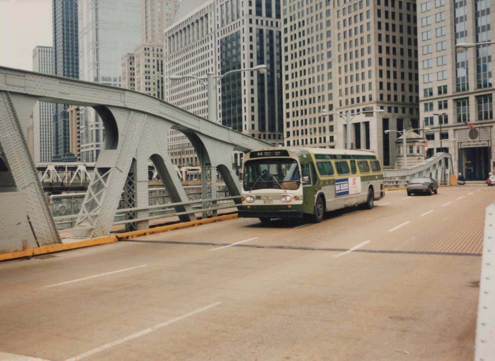 A green and white CTA bus, numbered 9101, drives on a steel bridge in a city with tall buildings in the background.