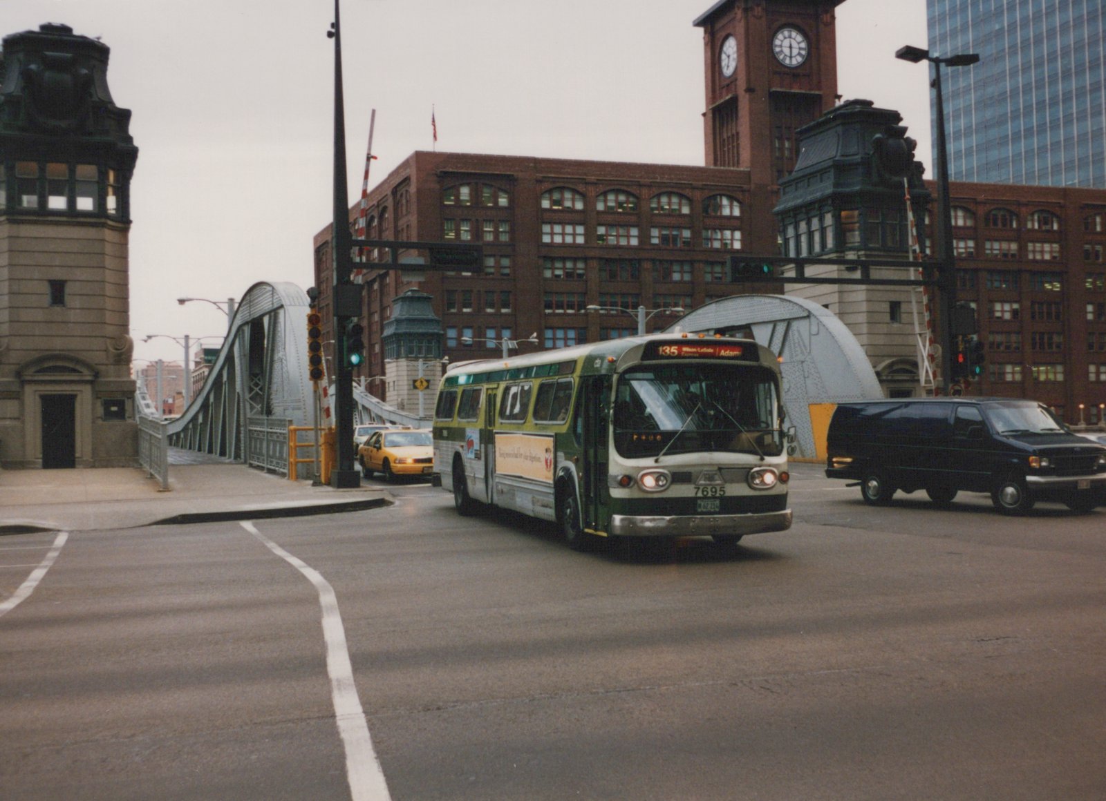 A green and cream CTA bus, number 7695, is turning on a city street next to a large steel bridge and a brick building with a clock tower.