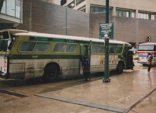 A green and white CTA bus, number 7607, is stopped on a wet city street near a sign for the Merchandise Mart as people board and walk by.