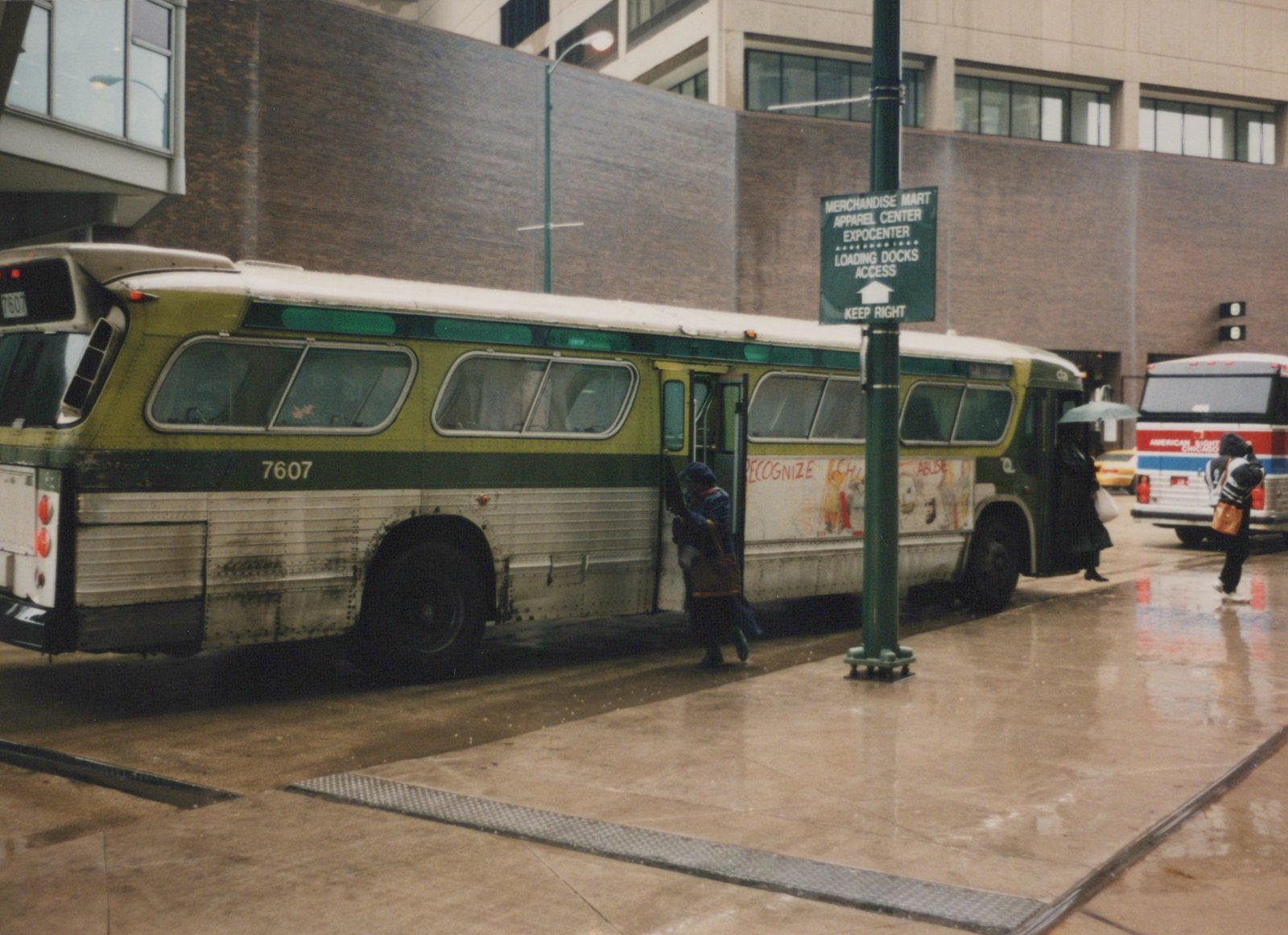 A green and white CTA bus, number 7607, is stopped on a wet city street near a sign for the Merchandise Mart as people board and walk by.