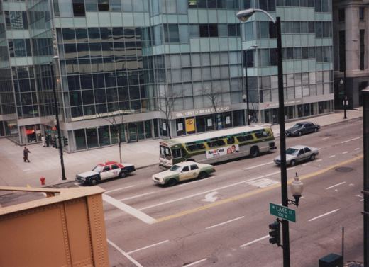 A green and cream CTA bus drives down a city street lined with glass buildings and taxis, with a street sign for W Lake St visible in the foreground.
