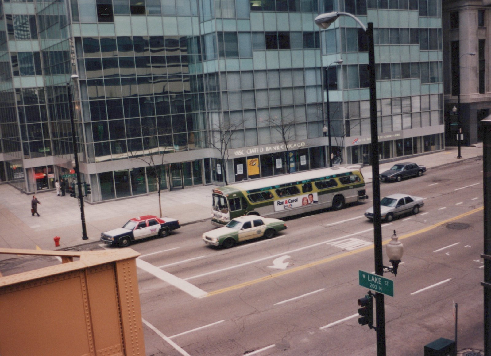 A green and cream CTA bus drives down a city street lined with glass buildings and taxis, with a street sign for W Lake St visible in the foreground.