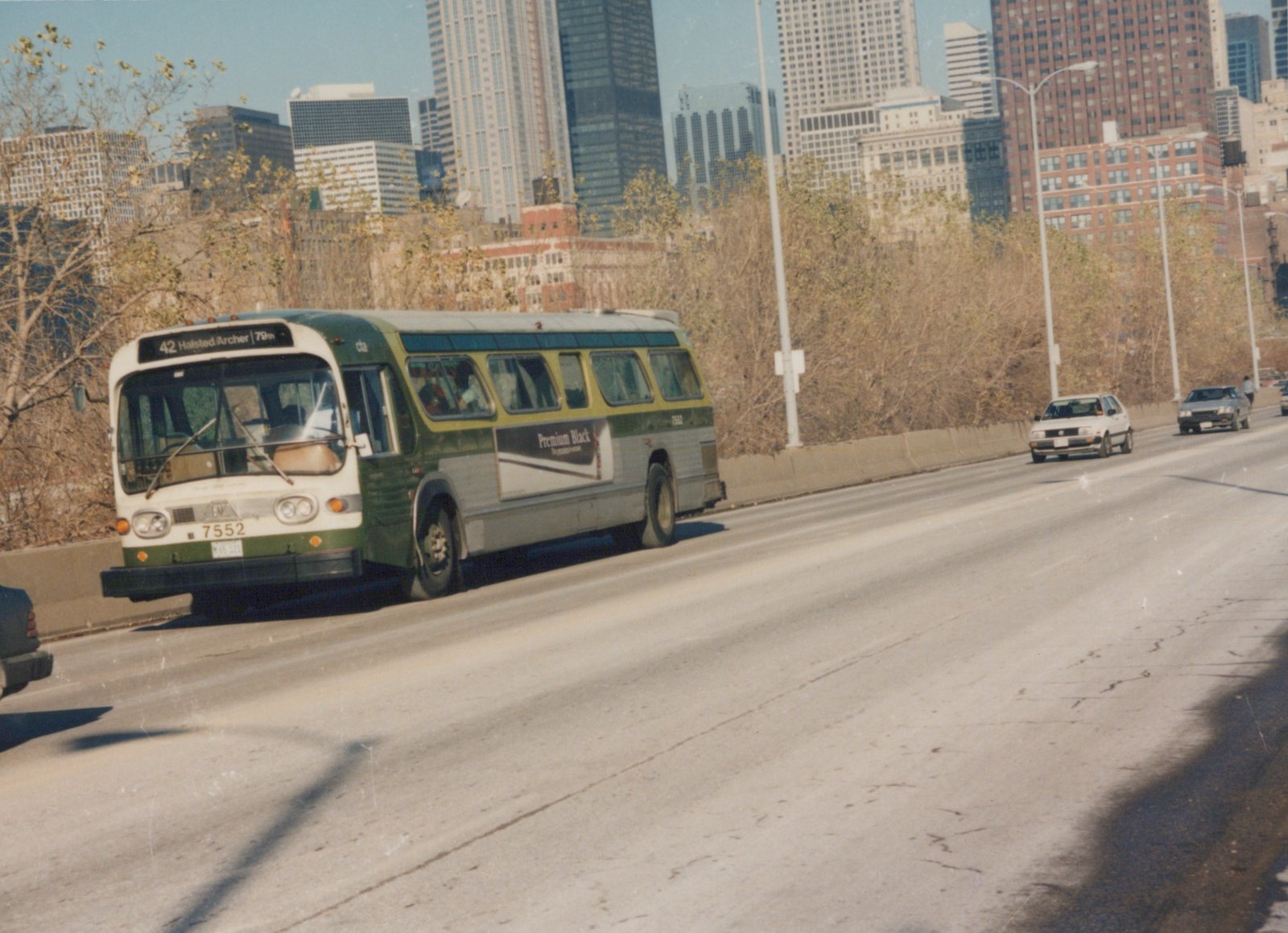 A green and cream CTA bus, number 7552, on a highway with tall buildings behind it on a sunny day.