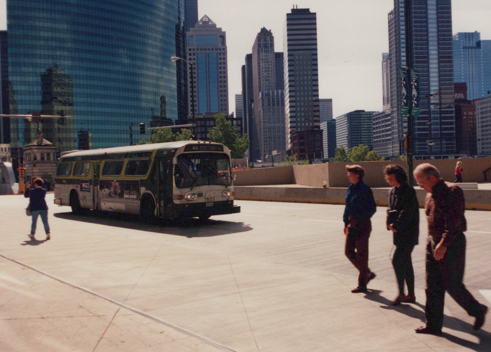 A white and green CTA bus, number 7503, is shown on a sunny day in a city setting with tall buildings behind it, while pedestrians walk nearby.