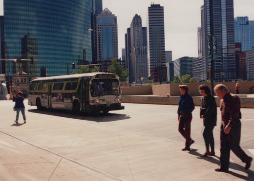 A white and green CTA bus, number 7503, is shown on a sunny day in a city setting with tall buildings behind it, while pedestrians walk nearby.