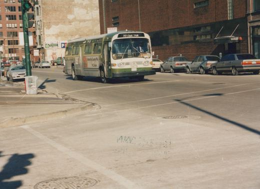 A green and cream Chicago Transit Authority bus is turning at an intersection on a sunny day, with several parked cars on the right and a traffic light on the left.