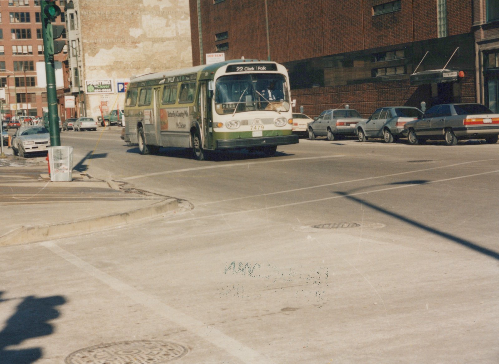 A green and cream Chicago Transit Authority bus is turning at an intersection on a sunny day, with several parked cars on the right and a traffic light on the left.