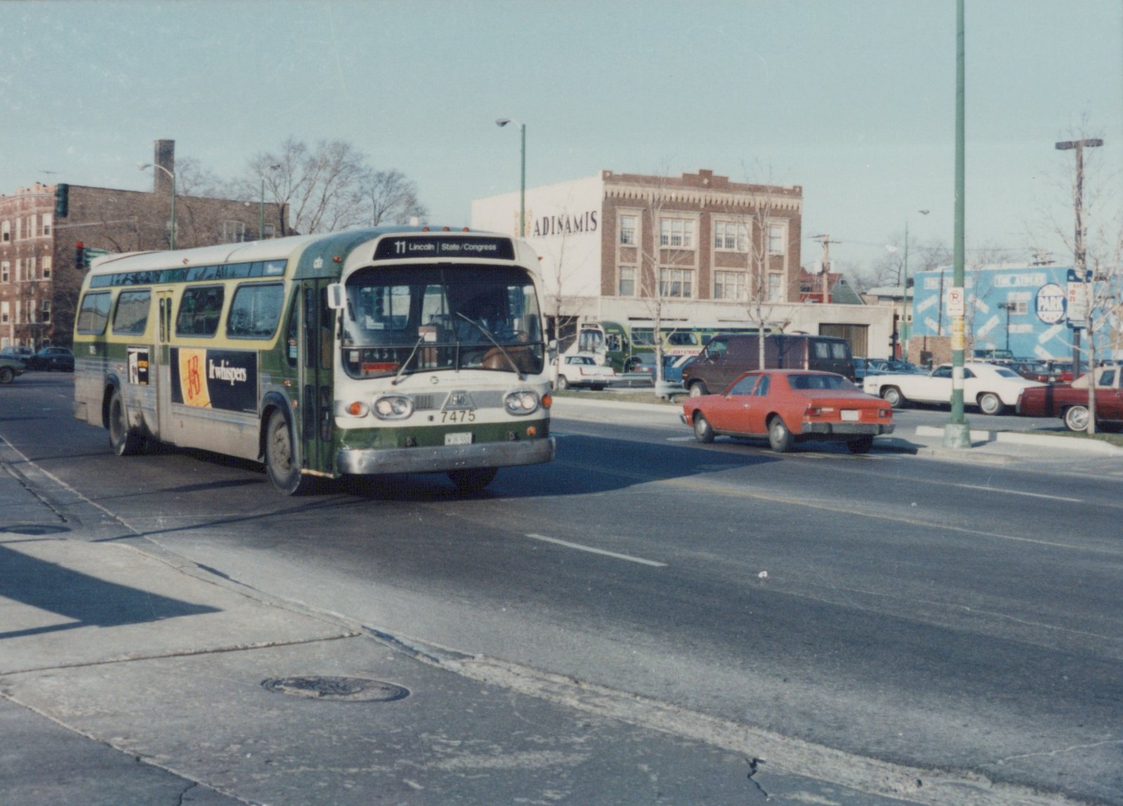 A green and cream CTA bus numbered 7475 drives down a city street in winter, with bare trees and other cars visible in the background.