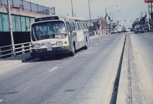 A green and cream motor bus with the number 7453 on its front bumper is driving down a multi-lane city street under a clear sky, with other cars and buildings visible in the background.