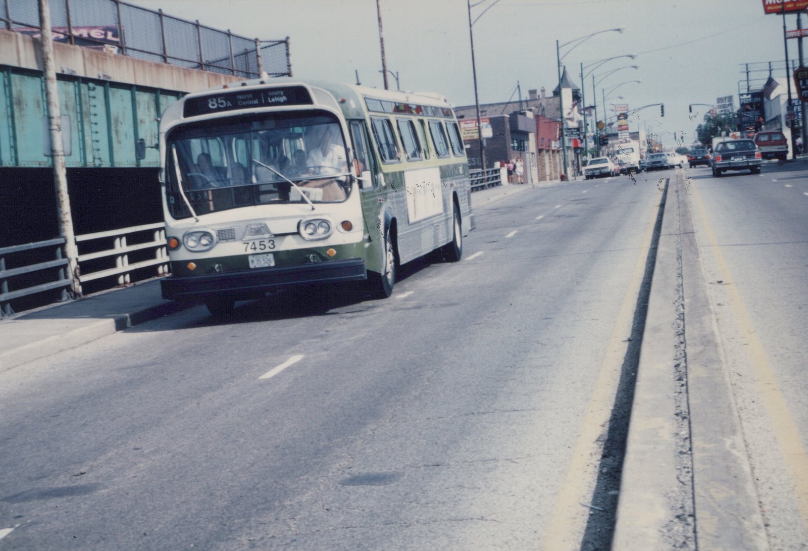 A green and cream motor bus with the number 7453 on its front bumper is driving down a multi-lane city street under a clear sky, with other cars and buildings visible in the background.