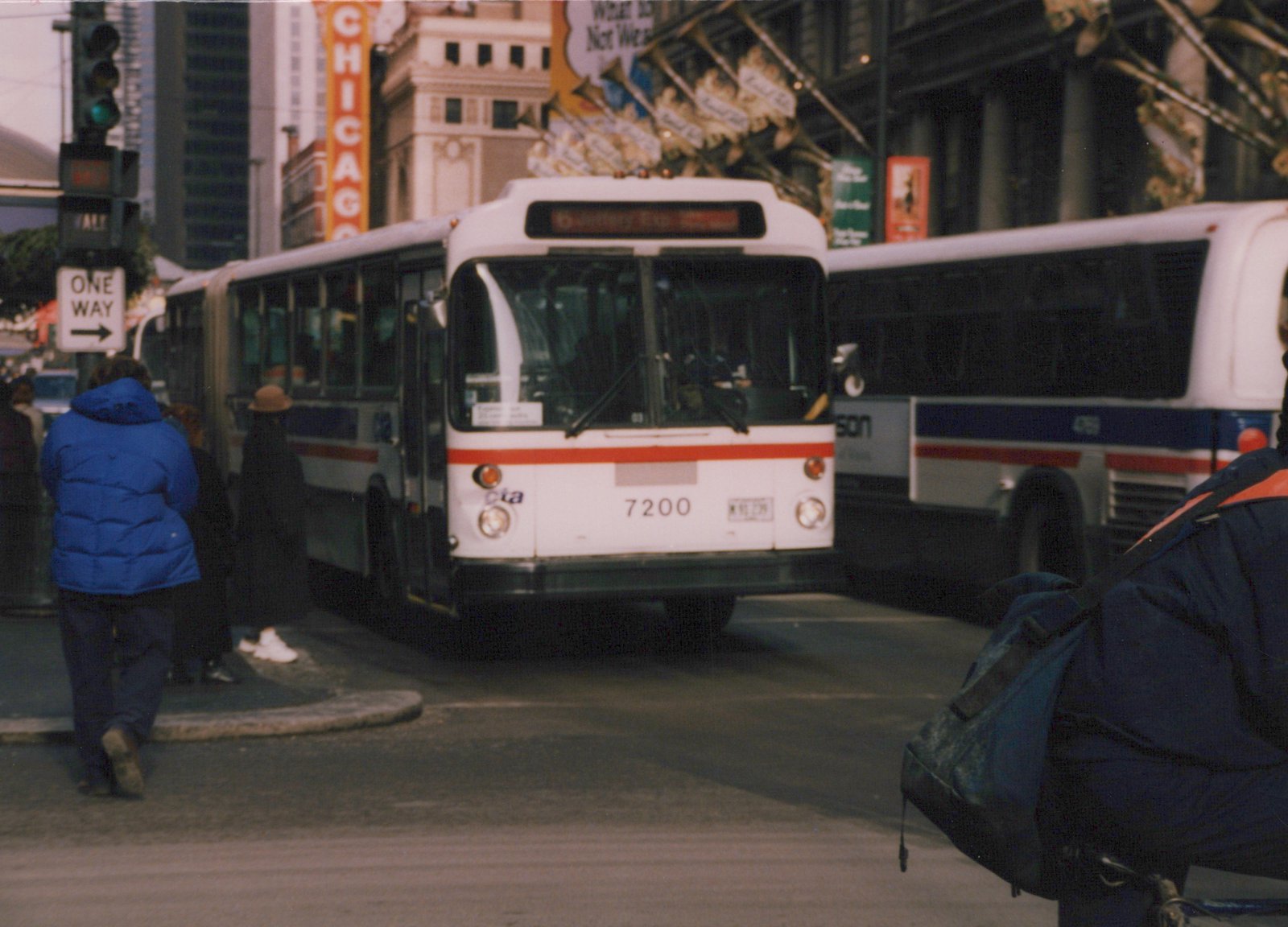 Front view of a white and red Chicago Transit Authority bus with the number 7200, stopped at an intersection with pedestrians and another bus visible.