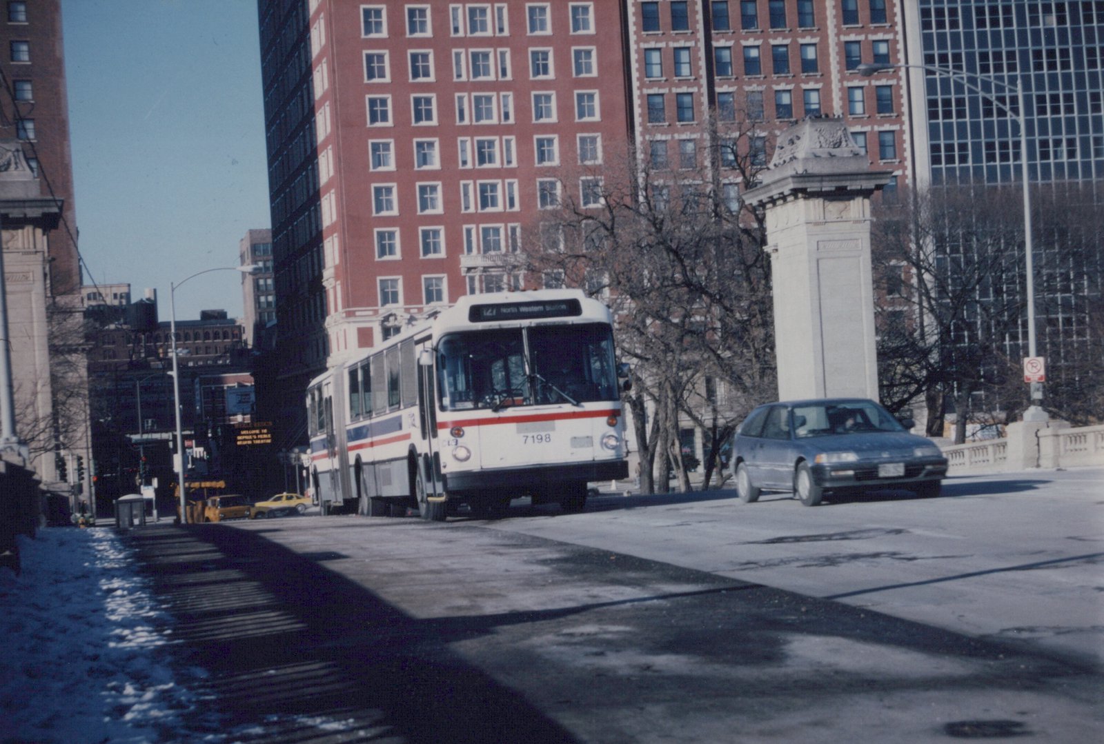 A white, red, and blue CTA bus numbered 7198 is driving on a city street with snow on the ground, past a blue car and tall buildings under a clear sky.