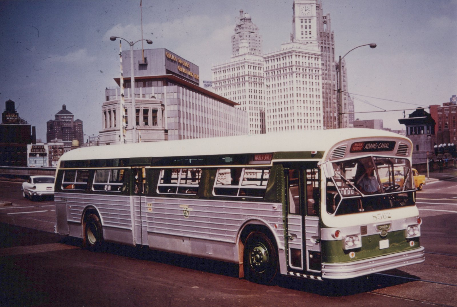 A silver and green Chicago Transit Authority bus, number 8562, is shown from a three-quarter front view on a city street, with tall buildings and a cloudy sky behind it.