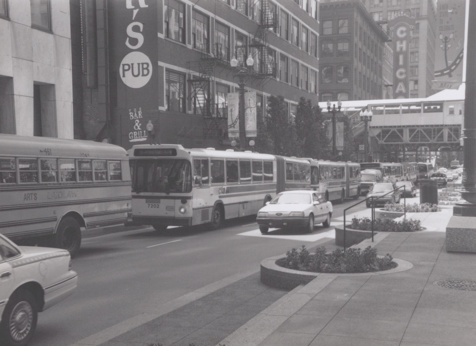 A black and white photo shows a CTA articulated bus on a city street lined with buildings, including a pub and the Chicago Theatre sign, with several cars and a school bus present.