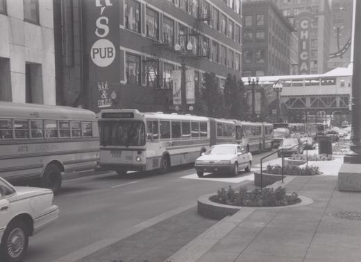 A black and white photo shows a CTA articulated bus on a city street lined with buildings, including a pub and the Chicago Theatre sign, with several cars and a school bus present.