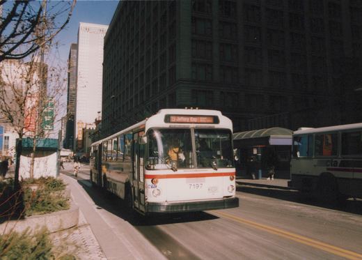 A white CTA bus with the number 7197 is driving down a city street past tall buildings, with another CTA bus and a cyclist visible in the background.