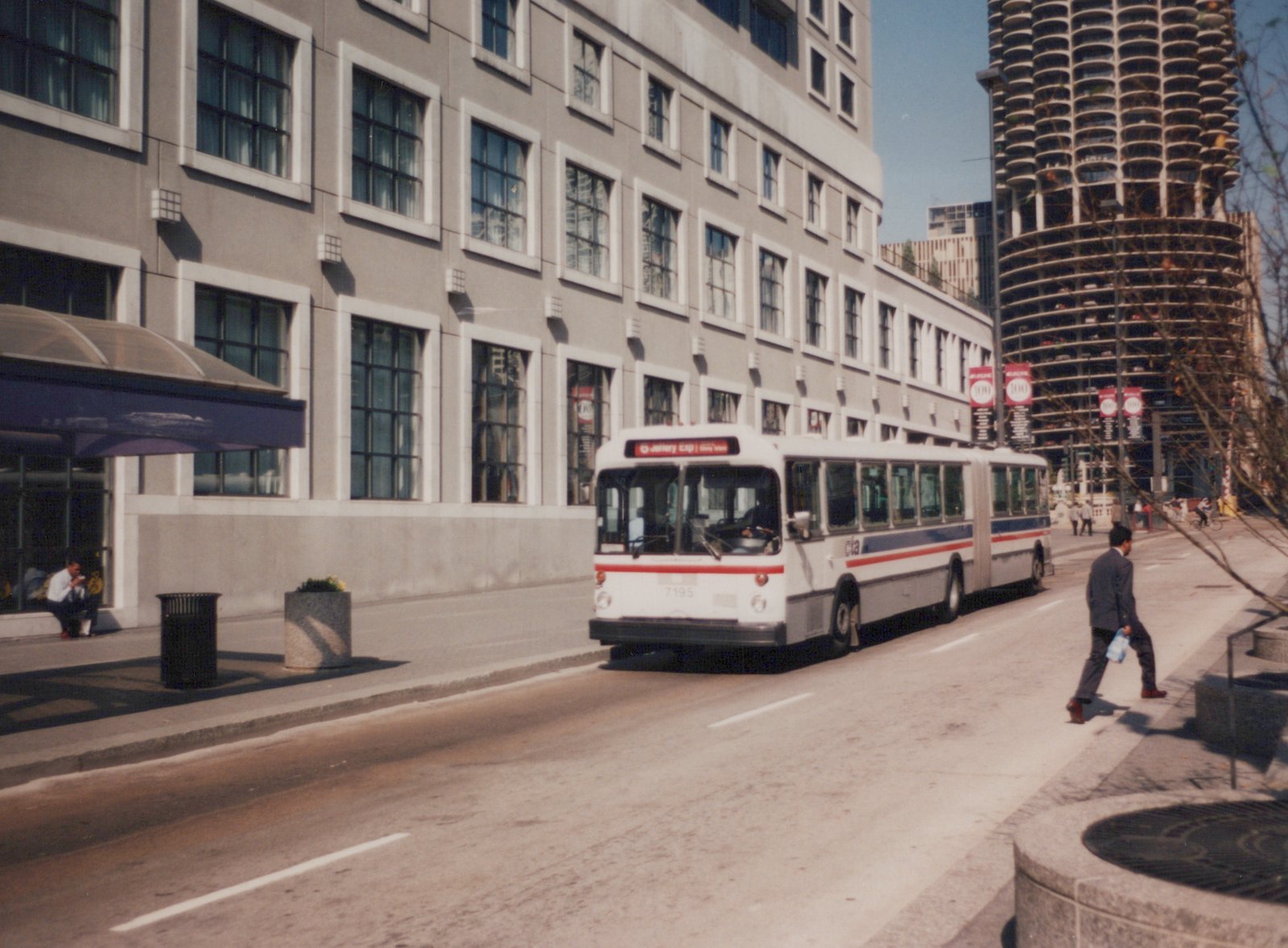 An articulated CTA bus, numbered 7195, drives down a city street with a pedestrian walking nearby and a distinctive round building in the background.