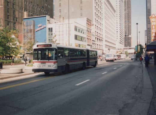 A white and blue CTA articulated bus, number 7182, is stopped on a Chicago street with tall buildings and pedestrians visible.