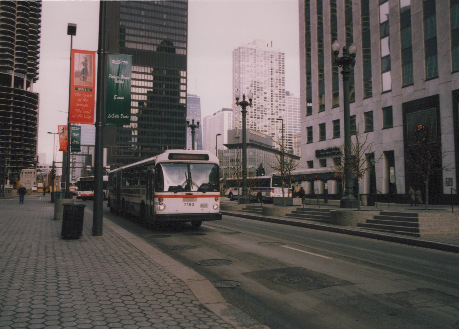 A white and orange CTA bus, number 7193, drives down a city street lined with tall buildings and banners, under an overcast sky.