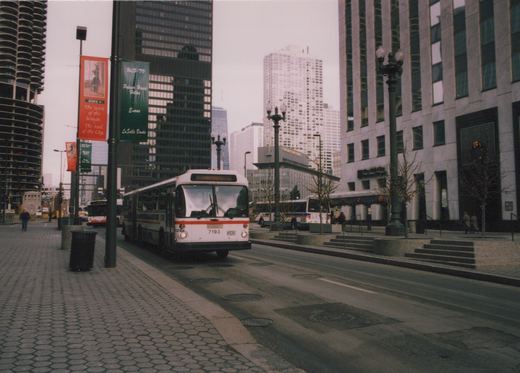 A white and orange CTA bus, number 7193, drives down a city street lined with tall buildings and banners, under an overcast sky.