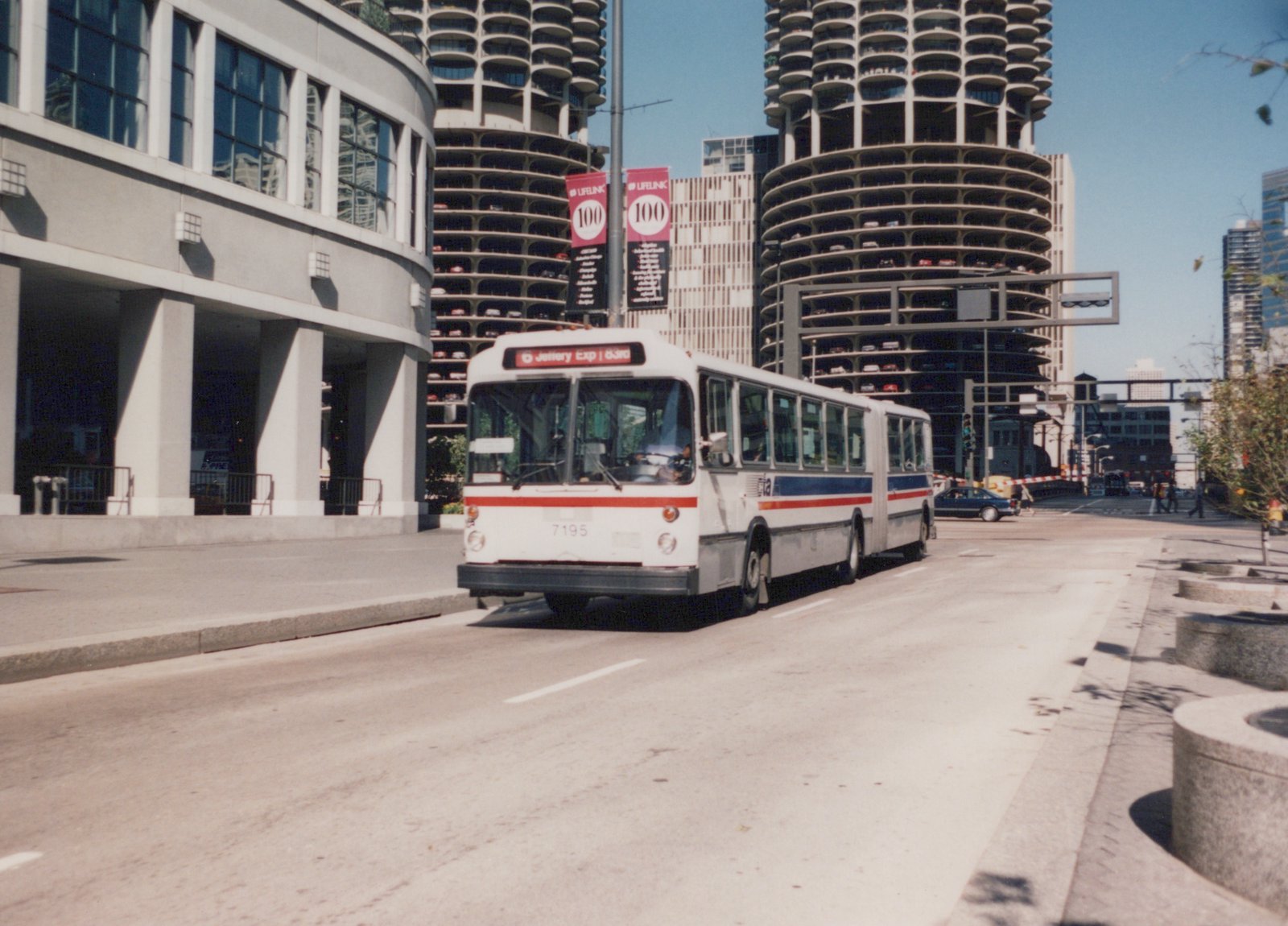 An articulated bus with the number 7195 and CTA lettering is parked on a city street, with the distinctive circular Marina City towers visible behind it under a clear blue sky.
