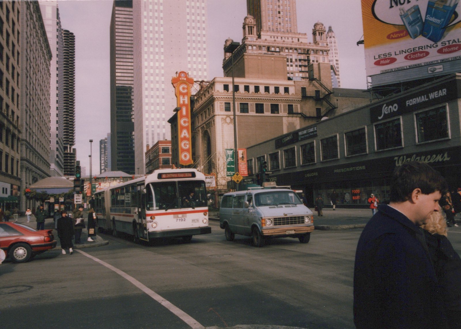 A white CTA bus with red and blue stripes drives down a Chicago street, with the Chicago Theater sign visible in the background and pedestrians on the sidewalks.