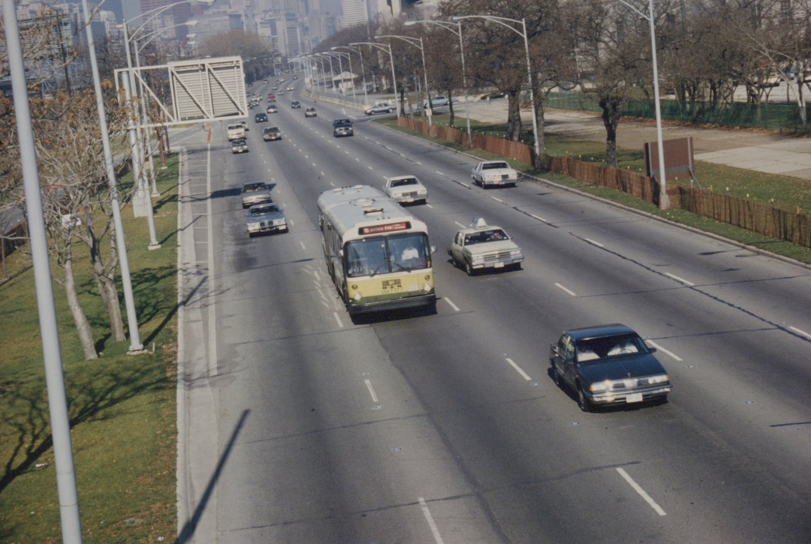 A yellow and white CTA motor bus numbered 7177 drives on a wide highway with several cars, including a taxi, under a clear sky with bare trees alongside the road.