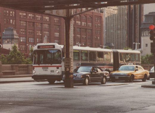 A white and red articulated city bus with the number 7169 on its front is stopped on a street next to a black sedan and a yellow taxi, with brick buildings and an elevated rail structure in the background.