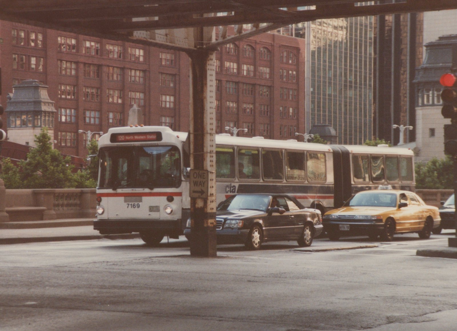 A white and red articulated city bus with the number 7169 on its front is stopped on a street next to a black sedan and a yellow taxi, with brick buildings and an elevated rail structure in the background.